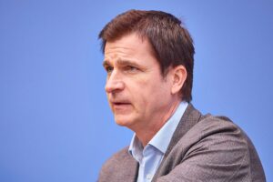 André Berghegger im Gespräch, hinter ihm ein urbanes Stadtbild, symbolisiert kommunale Herausforderungen.