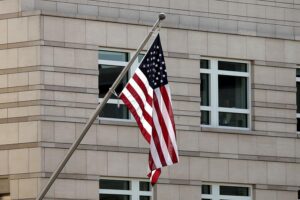 US-Flagge vor politischen Symbolen, die transatlantische Beziehungen und militärische Strategien thematisieren.