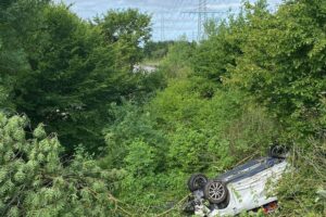 Wrack eines Fahrzeugs auf dem Dach, beschädigte Baumkrone im Hintergrund, Unfallstelle erkennbar.
