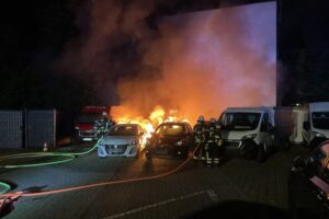 Feuerwehr löscht brennende PKW und Transporter, Einsatzkräfte schützen nahe Wohngebäude.