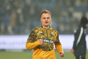 Jakob Lemmer in Dynamo-Dresden-Trikot, jubelnd auf dem Platz, umgeben von Fans und Teamkollegen.