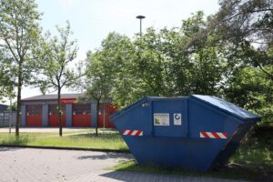 Ein Papiercontainer vor der Feuerwehr, umgeben von Müll, mit einem klaren Wochentagshinweis.