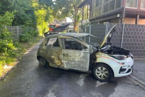 Ausgebrannter Pkw vor Wohn- und Geschäftshaus, Feuerwehr bei Löscharbeiten, Rauchentwicklung sichtbar.