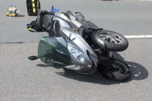 Verunfallter dreirädriger Roller auf der Straße, Zeugen helfen verletztem Fahrer. Flucht des Unfallverursachers.