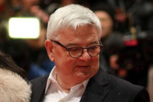 Joschka Fischer spricht, während Protestierende kritische Parolen skandieren und von der Polizei entfernt werden.