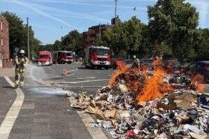 Brennendes Müllfahrzeug, Feuerwehr im Einsatz, Löscharbeiten, Straße gesperrt, Rauch und Einsatzkräfte.