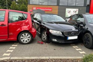 Ein Verkehrsunfall-Archivfoto: beteiligte Autos auf Supermarktparkplatz, Polizeiabsperrung, Verletzte nach Rückwärtsfahrt.