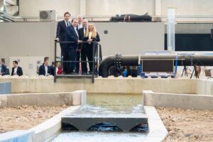 Wissenschaftler erklären Hochwasserschutzkonzepte, während Wasser am Brückenmodell staut. Politische Diskussion.