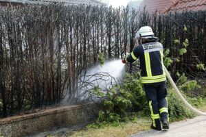 Einsatzkräfte löschen Heckenbrand, kontrollieren Glutnester mit Wärmebildkamera, warnen vor Brandgefahr.
