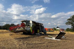 Ausgebrannter Mähdrescher auf einem Feld, Feuerwehr im Löscheinsatz, Brandrauch in der Luft.