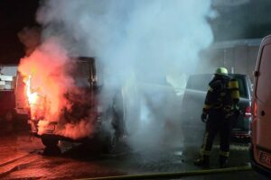 Brennende Kleintransporter, Feuerwehr im Einsatz, Nachtaufnahme, Rauch und Flammen, dramatische Szene.
