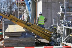 Bauarbeiter auf einer Baustelle, umgeben von Baustellenmaterialien und Bauzubehör, unter Normen-Diskussion.