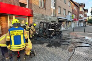 Eine im Vollbrand stehende Straßenkehrmaschine im Pulheimer Stadtzentrum vor einem Wohn- und Geschäftshaus.
